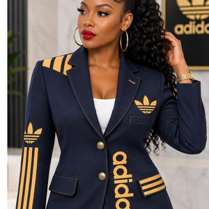 May include: A navy blue blazer featuring gold-colored Adidas branding, including the trefoil logo on the chest and shoulder, three stripes on the sleeve and pocket, and the word adidas printed vertically in gold along the front button closure.