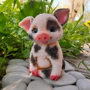 May include: A charming figurine of a piglet, featuring a pink snout, ears, and feet. The piglet has black spots on its white body and large, expressive eyes. It is sitting on a bed of white stones, surrounded by green foliage.