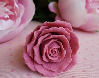 May include: A close-up of a dusty rose-colored rose with detailed petals. The rose is in focus, with two blurred pink flowers in the background. The background is a light pink fabric with small, darker pink polka dots.