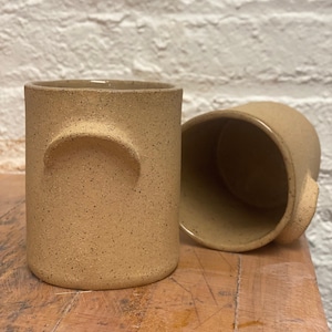 May include: Two beige ceramic mugs with a textured surface and a unique handle design. The mugs are sitting on a wooden surface.