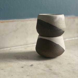 Two ceramic cups, each with a dark brown base and an off-white top, are stacked on top of each other on a white marble surface.