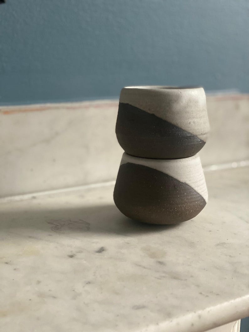 Two ceramic cups, each with a dark brown base and an off-white top, are stacked on top of each other on a white marble surface.
