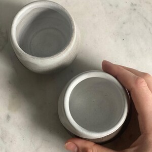 A hand holds one ceramic cup, showing its off-white interior, next to another similar cup on a white marble surface.