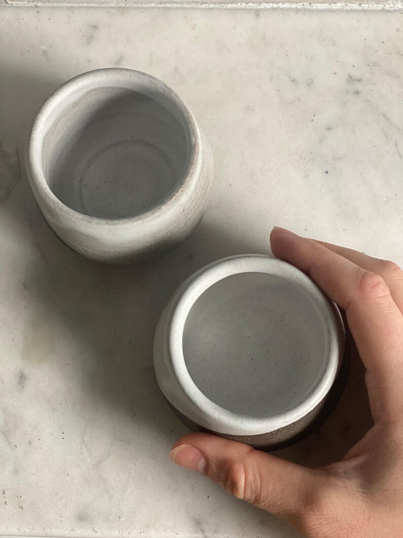 A hand holds one ceramic cup, showing its off-white interior, next to another similar cup on a white marble surface.