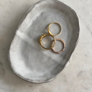 May include: A white ceramic ring dish with a textured surface. Four gold rings are resting in the dish.