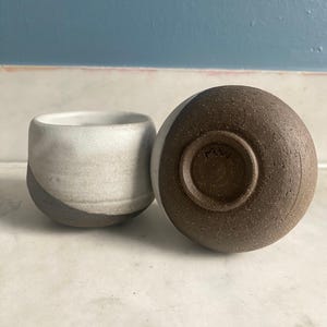 One ceramic cup stands upright while another lies on its side, revealing its dark brown, textured base with a visible maker's mark.