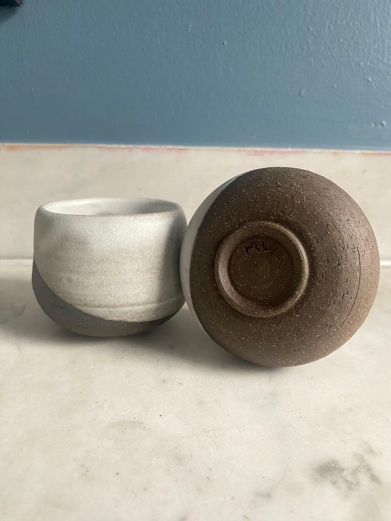 One ceramic cup stands upright while another lies on its side, revealing its dark brown, textured base with a visible maker's mark.
