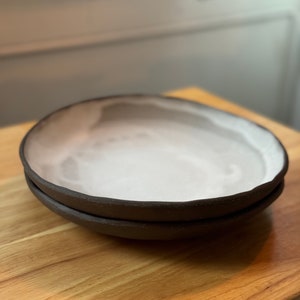 Puede incluir: Dos platos de cerámica hechos a mano apilados sobre una superficie de madera. Los platos tienen un borde marrón oscuro y un interior blanco con un patrón sutil y orgánico. Los platos son redondos y parecen poco profundos.