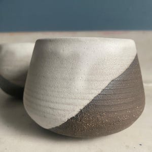 A close-up shows a single ceramic cup with a textured off-white upper portion and a dark brown, ridged lower portion, sitting on a light surface.