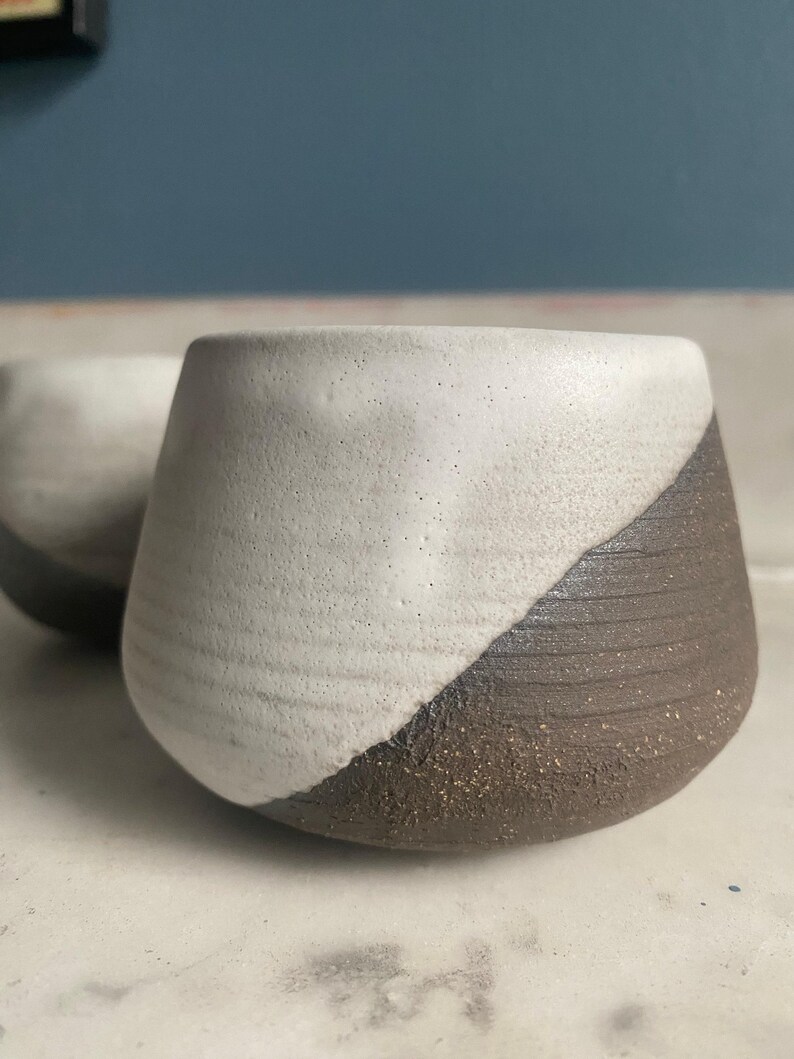 A close-up shows a single ceramic cup with a textured off-white upper portion and a dark brown, ridged lower portion, sitting on a light surface.