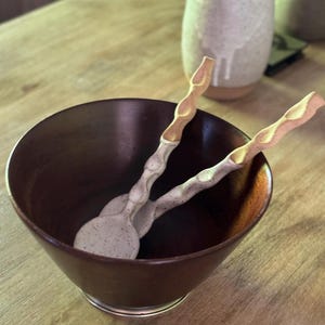 May include: A dark brown ceramic bowl with two matching serving spoons. The spoons have a textured, light-coloured handle and a speckled, light-coloured spoon head. The bowl sits on a wooden surface, with a decorative vase in the background.