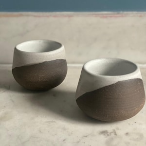 Two handmade ceramic espresso cups, each with a dark brown base and an off-white top, sit on a white marble surface against a blue wall.
