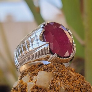 May include: A silver ring with a large, oval-shaped ruby gemstone. The ring has a textured band with a geometric design.