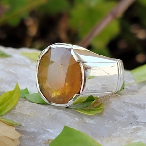 May include: A silver ring with a large, oval-shaped, yellow gemstone set in a bezel setting. The ring has a thick band with a polished finish.