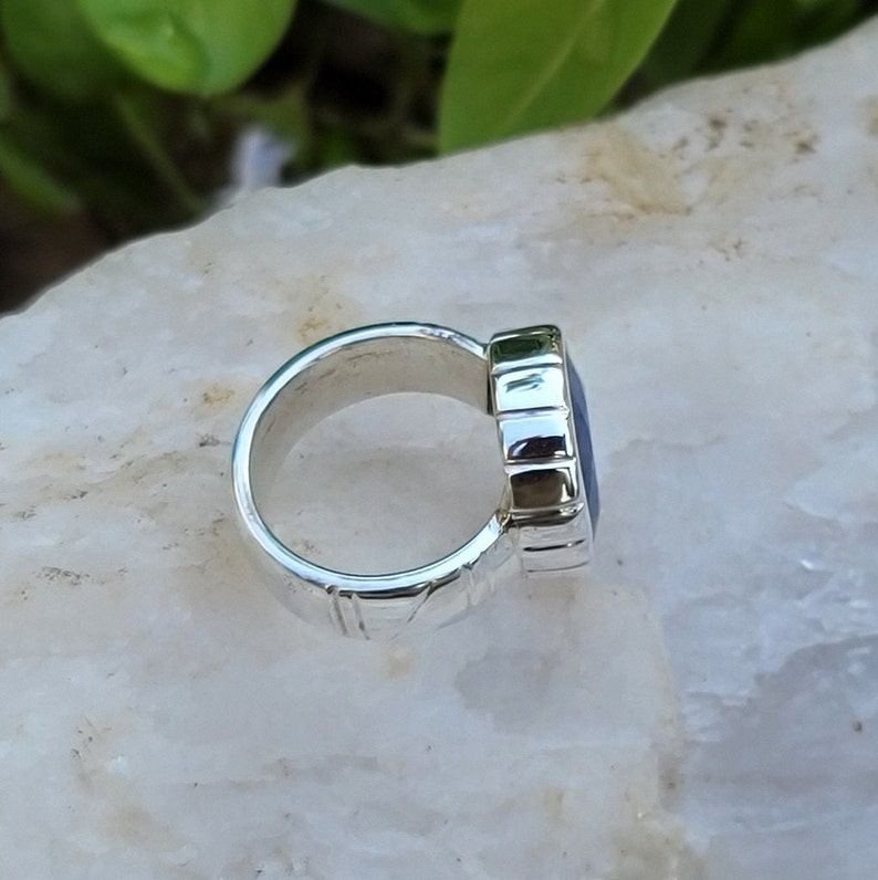 May include: A silver ring with a dark blue gemstone set in a textured bezel. The ring is on a white background.