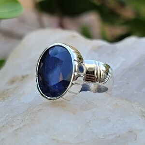May include: A silver ring with a large, oval-shaped blue sapphire gemstone. The ring has a detailed band with a textured design.