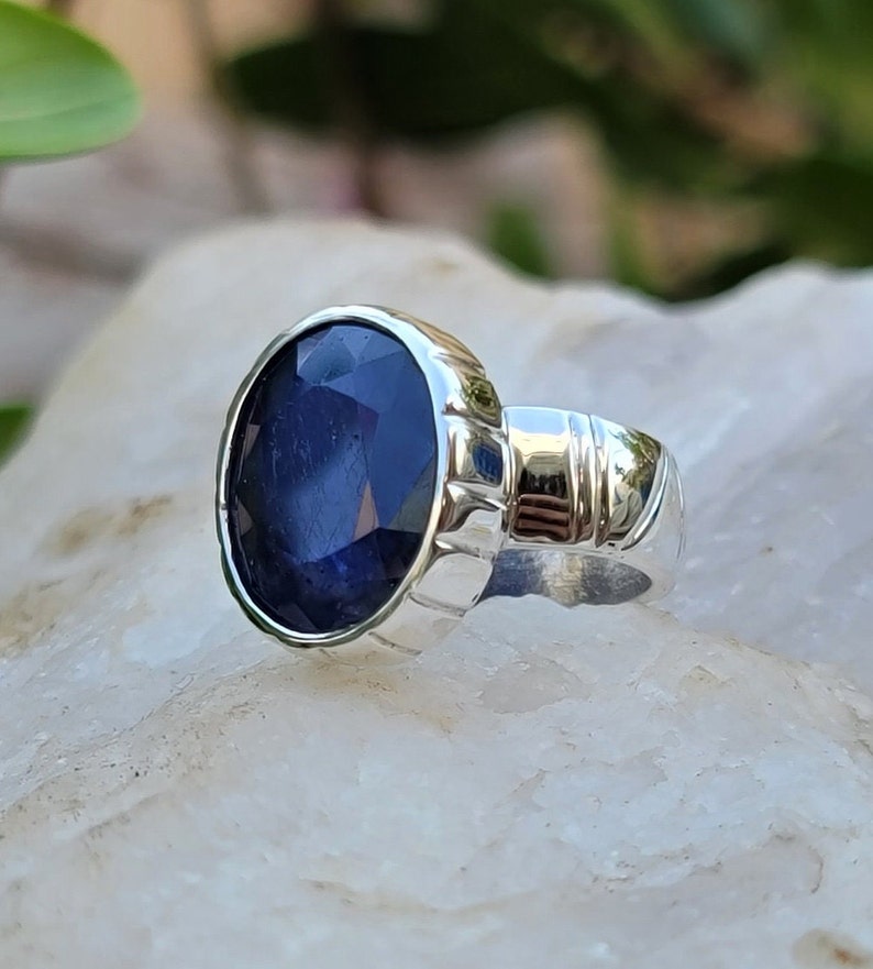 May include: A silver ring with a large, oval-shaped blue sapphire gemstone. The ring has a detailed band with a textured design.