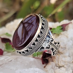 May include: A silver ring with an oval-shaped, dark red gemstone set in a detailed, ornate silver band.