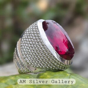 Peut inclure: Une bague en argent avec une grande pierre précieuse ovale de couleur rouge foncé. La bande de la bague est texturée avec de petites perles rondes.