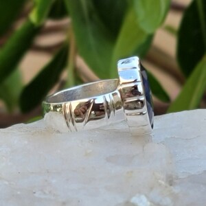 May include: A silver ring with a dark blue gemstone set in a faceted bezel. The band is textured with a repeating pattern.