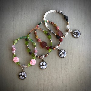 May include: Four layered bracelets featuring a variety of coloured beads and charms. The charms include a deer, a rabbit, a flower, and an owl. The bracelets are made with lava rock, wood, and glass beads.