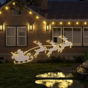 Op de afbeelding: Een wit silhouet van Sinterklaas in een slee getrokken door rendieren, verlicht met witte lampjes tegen een bakstenen huis met kerstverlichting.