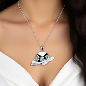 Puede incluir: Un collar de plata con un colgante de tortuga. El colgante tiene un caparazón blanco con detalles negros e iridiscentes de concha de abulón. La cabeza y las aletas de la tortuga son blancas.