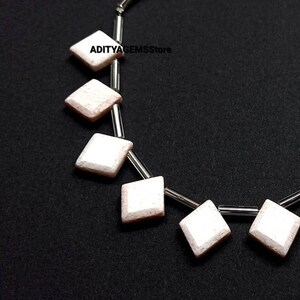 May include: A strand of white and pink gemstone beads strung on a silver chain. The beads are square-shaped with a diamond-like cut.
