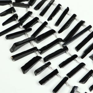 May include: Close-up of a necklace featuring glossy black rectangular beads strung together with silver-colored wire. The beads are of varying lengths and shapes, creating a unique, geometric design. The background is plain white.