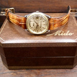 Puede incluir: Un reloj Mido vintage con esfera dorada y correa de cuero marrón con estampado de cocodrilo. El reloj descansa sobre una caja marrón con el nombre de la marca "Mido" en letras doradas. La caja tiene un patrón de greca decorativo.