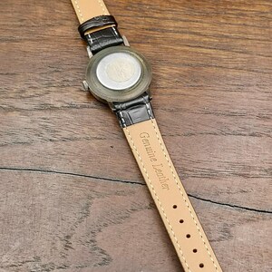 May include: A wristwatch with a black crocodile-textured strap and a silver-tone metal case back. The inner side of the strap is tan, featuring the embossed text Genuine Leather and the number 16 near the end, with a gold-tone metal buckle.