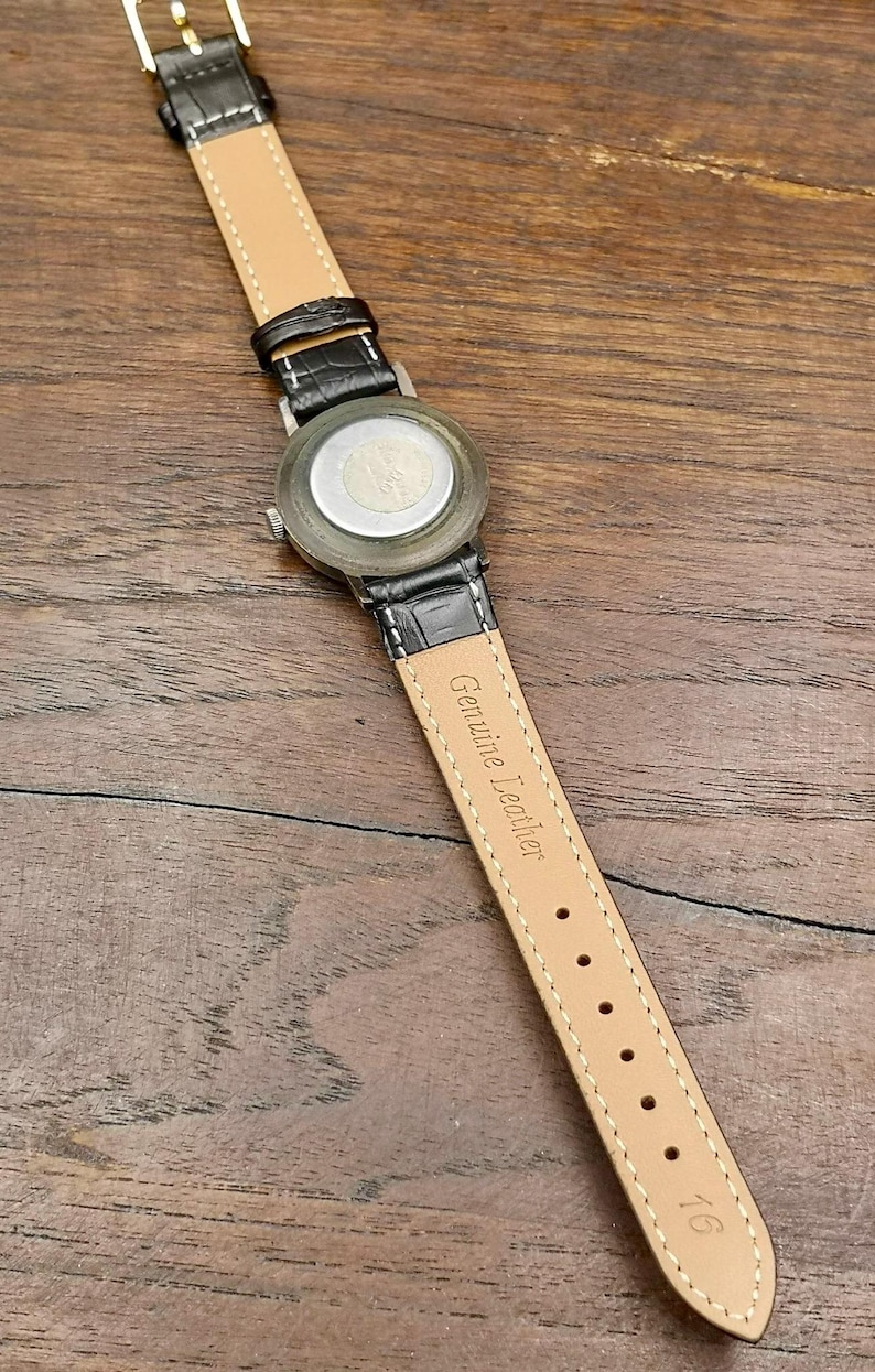 May include: A wristwatch with a black crocodile-textured strap and a silver-tone metal case back. The inner side of the strap is tan, featuring the embossed text Genuine Leather and the number 16 near the end, with a gold-tone metal buckle.