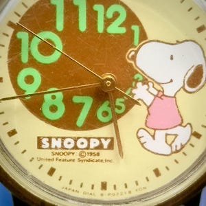May include: A round analog watch face with a cream background, featuring a brown circle in the center with large, light green numbers from 6 to 12. Snoopy is depicted on the right wearing a pink shirt. Text includes SNOOPY, 1958 United Feature Syndicate, Inc.