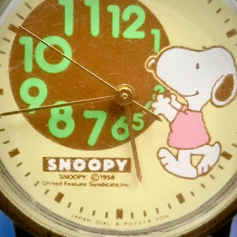 May include: A round analog watch face with a cream background, featuring a brown circle in the center with large, light green numbers from 6 to 12. Snoopy is depicted on the right wearing a pink shirt. Text includes SNOOPY, 1958 United Feature Syndicate, Inc.