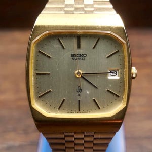 Op de afbeelding: Een goudkleurig Seiko Quartz horloge met een rechthoekige wijzerplaat en bijpassende schakelband. De wijzerplaat van het horloge is lichtgoudkleurig met zwarte wijzers en markeringen. De datum wordt weergegeven op 3 uur. Het horloge is gemarkeerd met "SEIKO QUARTZ" en "JAPAN 7822-5000 T".