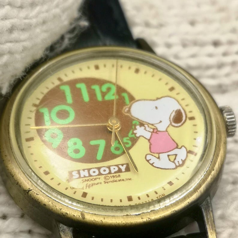 May include: A vintage-style wristwatch with a round brass-toned case and a cream-colored dial. The face features a brown center circle with large green numerals, a graphic of Snoopy wearing a pink shirt, and the text SNOOPY 1958 United Feature Syndicate, Inc.