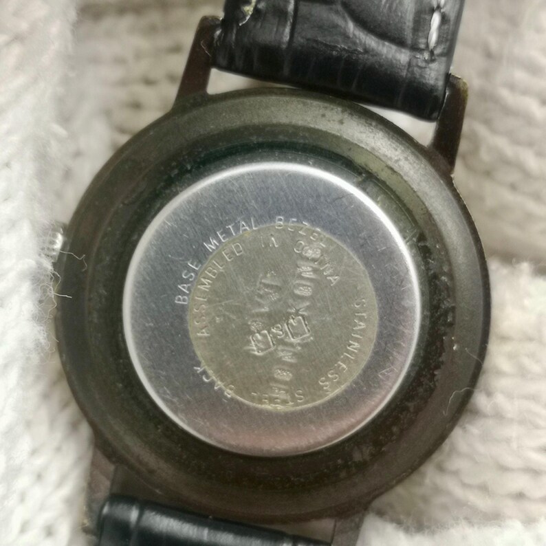 May include: The back of a round wristwatch with a black leather strap. The silver-toned metal case back features text that reads Base Metal Bezel, Assembled in China, and Stainless Steel Back. The watch casing is dark-toned and circular in shape.