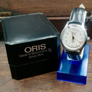 Puede incluir: Un reloj Oris plateado con esfera blanca y correa de cuero negro, exhibido sobre un soporte azul. La esfera del reloj tiene números y agujas negros. Una caja negra con el logotipo de Oris y "Made in Switzerland Since 1904" está junto al reloj.