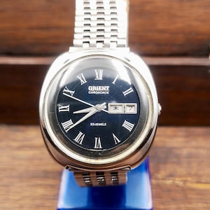 May include: A silver-tone Orient Chronoace wristwatch with a dark blue circular dial, silver Roman numeral hour markers, and a metal link bracelet. The watch face displays the text Orient Chronoace, 23 Jewels, and a day-date window with a Japanese character.