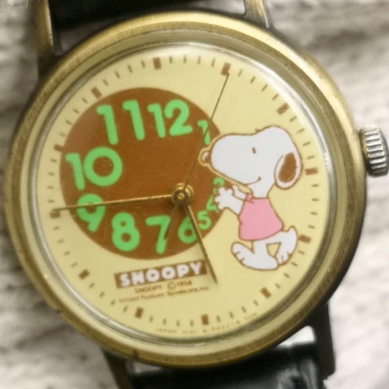 May include: A round analog wristwatch with a brass-toned metal case and a black leather strap. The cream-colored dial features a graphic of Snoopy wearing a pink shirt, green numerals from 7 to 12, and the text SNOOPY 1958 United Feature Syndicate, Inc.