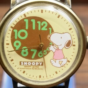 May include: A round analog wristwatch with a cream-colored face featuring a brown circle containing large green numbers 7 through 12. Snoopy is depicted in a pink shirt on the right side. Text includes SNOOPY, 1958 United Feature Syndicate, Inc.