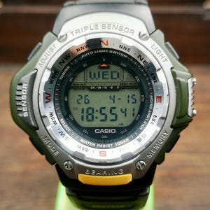 Peut inclure: Montre numérique Casio avec bracelet noir et vert olive. L'écran affiche la date, l'heure et le jour de la semaine. Fonctions : boussole, altimètre et résistance à l'eau 10 bar.