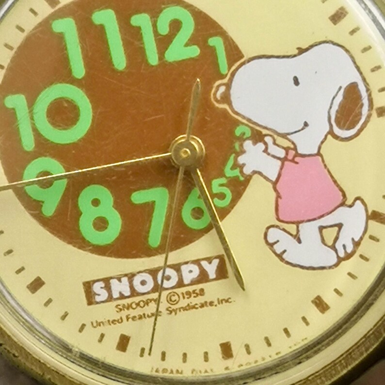 May include: A round analog watch face featuring a light yellow background with a brown center circle. Bright green numbers 7 through 12 are displayed. A white Snoopy character wearing a pink shirt is printed on the right. Text reads SNOOPY &copy;1958 United Feature Syndicate, Inc.