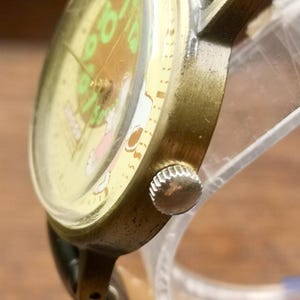 May include: A wristwatch with a metallic gold-toned case and a silver-colored crown on the side. The watch face features a light yellow background with green numerals and a small graphic illustration. It includes a black strap attached to the lugs.