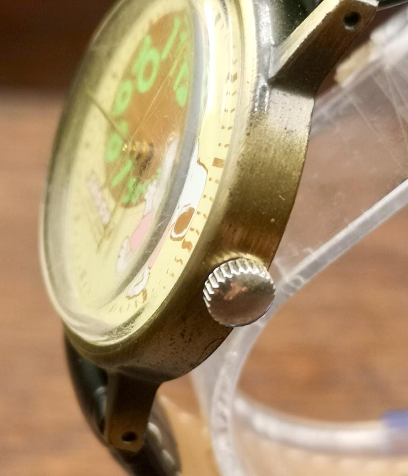 May include: A wristwatch with a metallic gold-toned case and a silver-colored crown on the side. The watch face features a light yellow background with green numerals and a small graphic illustration. It includes a black strap attached to the lugs.