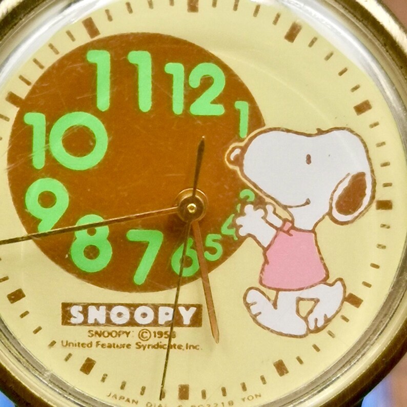 May include: A round analog watch face featuring a cream-colored background with a brown circular center. Bright green numerals 7 through 12 are displayed. A white Snoopy character wearing a pink shirt stands on the right. Text reads SNOOPY 1958.
