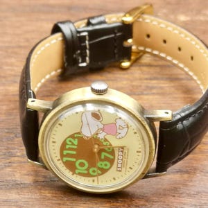 May include: A wristwatch with a round, antique-gold tone metal case and a black leather strap. The watch face features a cream background with a graphic of Snoopy, large green numerals, and the text "SNOOPY &copy;1958 United Feature Syndicate, Inc."