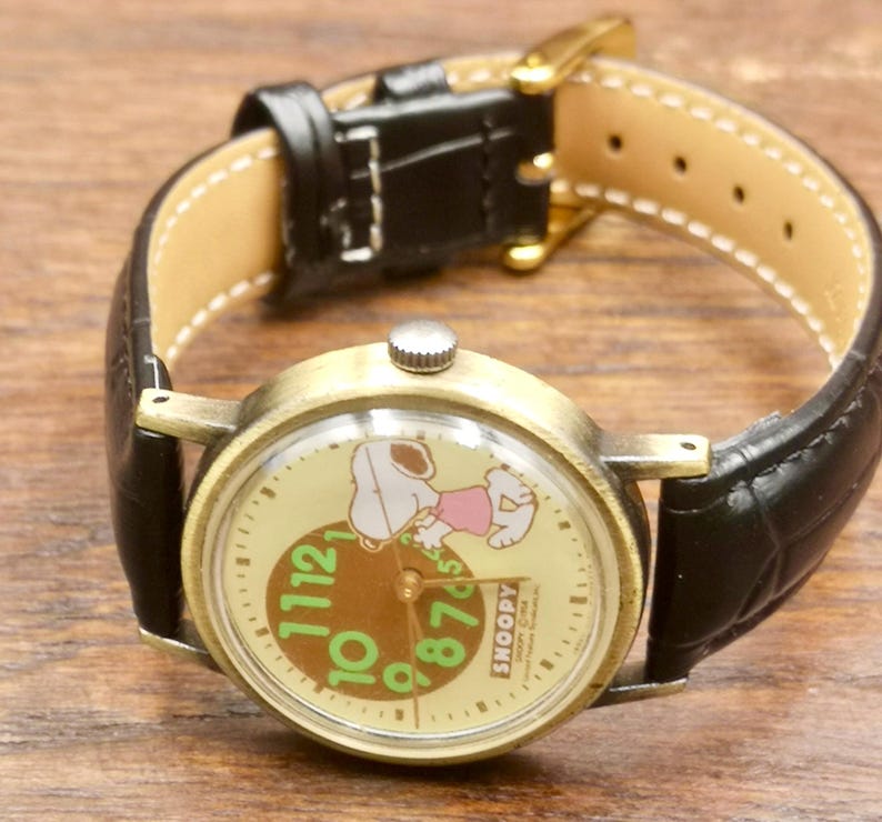 May include: A wristwatch with a round, antique-gold tone metal case and a black leather strap. The watch face features a cream background with a graphic of Snoopy, large green numerals, and the text "SNOOPY &copy;1958 United Feature Syndicate, Inc."