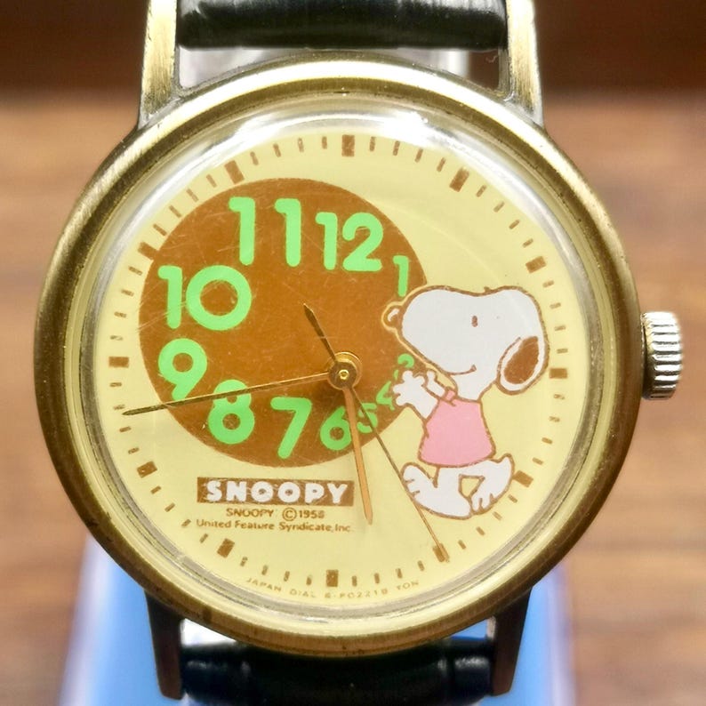 May include: A round analog wristwatch with a brass-toned metal case and a black leather strap. The cream-colored dial features a large brown circle with bright green numbers 7 through 12, a Snoopy character illustration, and the text SNOOPY 1958.
