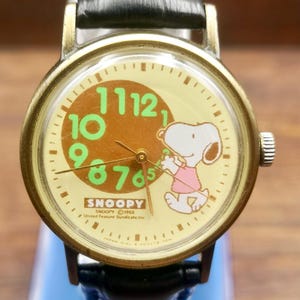 May include: A wristwatch with a round brass-toned case and a black leather strap. The cream-colored dial features a brown circle with large green numbers, a graphic of Snoopy wearing a pink shirt, and the text SNOOPY 1958 United Feature Syndicate, Inc.